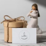 Caja de regalo de papel kraft decorada con una cuerda, junto a una tarjeta con el logo de Llamarena y una figura decorativa en el fondo. Ideal para presentaciones de productos artesanales.