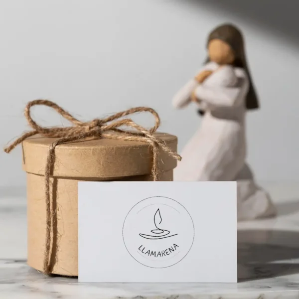 Caja de regalo de papel kraft decorada con una cuerda, junto a una tarjeta con el logo de Llamarena y una figura decorativa en el fondo. Ideal para presentaciones de productos artesanales.