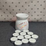 Difusor de cerámica blanca con diseño de hojas, acompañado de velas tipo té. Fondo textil con estampado de flores. Ideal para aromaterapia y ambientación.