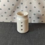Vaso de cerámica blanca con tres agujeros decorativos, colocado sobre un fondo de tela gris con estampado floral. Ideal para decoración o como soporte para velas.