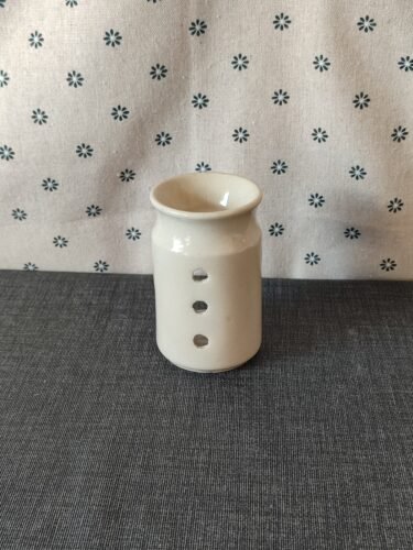 Vaso de cerámica blanca con tres agujeros decorativos, colocado sobre un fondo de tela gris con estampado floral. Ideal para decoración o como soporte para velas.
