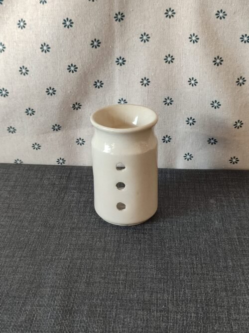 Vaso de cerámica blanca con tres agujeros decorativos, colocado sobre un fondo de tela gris con estampado floral. Ideal para decoración o como soporte para velas.