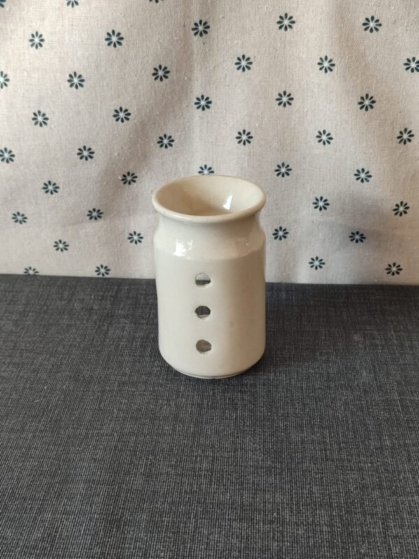 Vaso de cerámica blanca con tres agujeros decorativos, colocado sobre un fondo de tela gris con estampado floral. Ideal para decoración o como soporte para velas.