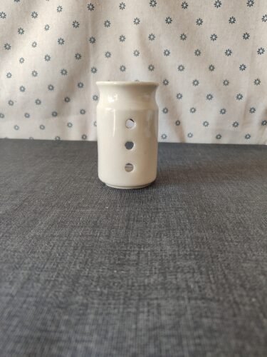 Vaso de cerámica blanca con tres agujeros decorativos, ubicado sobre una mesa gris, con fondo de tela blanca con estampado de flores azules. Ideal para decoración o uso como portavelas.