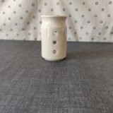Vaso de cerámica blanca con tres agujeros decorativos, ubicado sobre una mesa gris, con fondo de tela blanca con estampado de flores azules. Ideal para decoración o uso como portavelas.