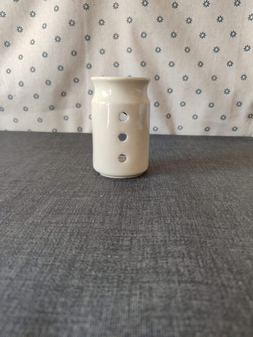 Vaso de cerámica blanca con tres agujeros decorativos, ubicado sobre una mesa gris, con fondo de tela blanca con estampado de flores azules. Ideal para decoración o uso como portavelas.