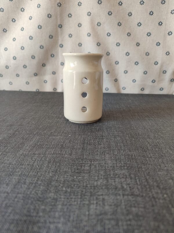 Vaso de cerámica blanca con tres agujeros decorativos, ubicado sobre una mesa gris, con fondo de tela blanca con estampado de flores azules. Ideal para decoración o uso como portavelas.