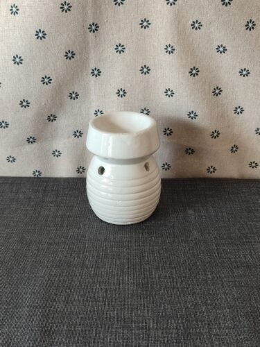 Cerámica blanca decorativa, con diseño estriado y orificios, ubicada sobre una superficie gris, frente a un fondo de tela con estampado de flores. Ideal para aromaterapia o como elemento decorativo.