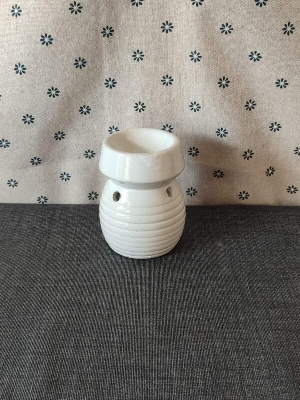 Cerámica blanca decorativa, con diseño estriado y orificios, ubicada sobre una superficie gris, frente a un fondo de tela con estampado de flores. Ideal para aromaterapia o como elemento decorativo.