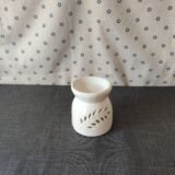 Difusor de cerámica blanca con diseño de hoja, colocado sobre una superficie gris, con fondo de tela con estampado de flores. Ideal para aromatizar espacios.