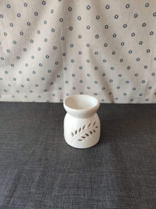 Difusor de cerámica blanca con diseño de hoja, colocado sobre una superficie gris, con fondo de tela con estampado de flores. Ideal para aromatizar espacios.