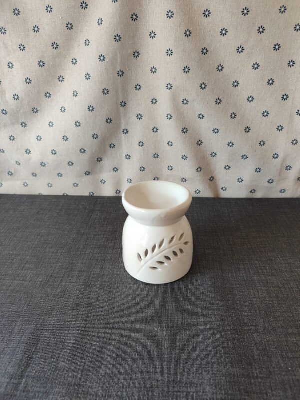 Difusor de cerámica blanca con diseño de hoja, colocado sobre una superficie gris, con fondo de tela con estampado de flores. Ideal para aromatizar espacios.