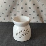 Difusor de cerámica blanca con diseño de hojas, ideal para aromatizar espacios. Fondo textil con estampado de flores.