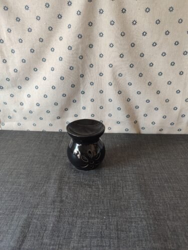 Jarrón de cerámica negra con diseño floral, sobre una mesa gris y fondo de tela blanca con estampado de flores azules. Ideal para decoración del hogar.