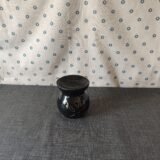 Jarrón de cerámica negra con diseño floral, sobre una mesa gris y fondo de tela blanca con estampado de flores azules. Ideal para decoración del hogar.