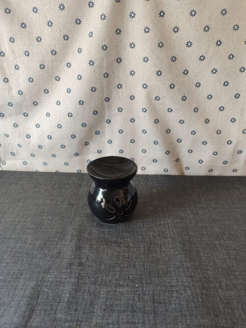 Jarrón de cerámica negra con diseño floral, sobre una mesa gris y fondo de tela blanca con estampado de flores azules. Ideal para decoración del hogar.