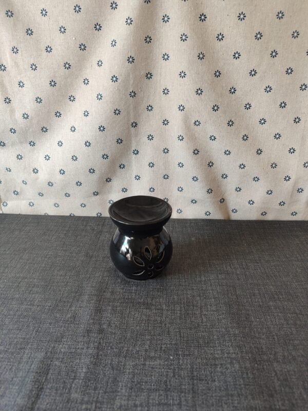 Jarrón de cerámica negra con diseño floral, sobre una mesa gris y fondo de tela blanca con estampado de flores azules. Ideal para decoración del hogar.