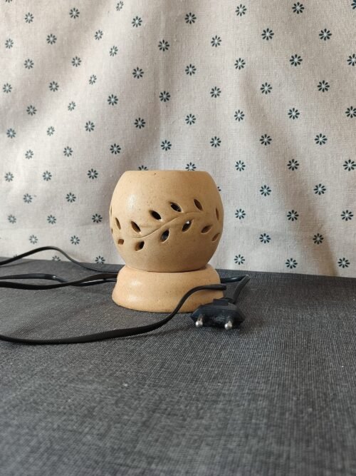 Lámpara de cerámica con diseño de hojas, conectada a un cable eléctrico, sobre una superficie textil gris. Ideal para decoración y aromaterapia.