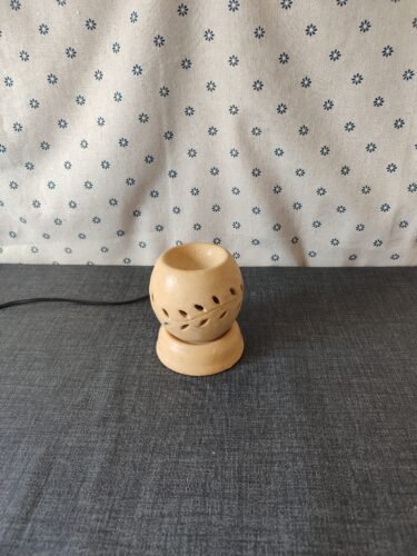 Lámpara de cerámica decorativa con diseño de hojas, sobre una superficie gris, con fondo de tela blanca estampada. Ideal para aromaterapia y decoración del hogar.