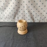 Lámpara de cerámica decorativa con diseño de hojas, sobre una superficie gris, con fondo de tela blanca estampada. Ideal para aromaterapia y decoración del hogar.