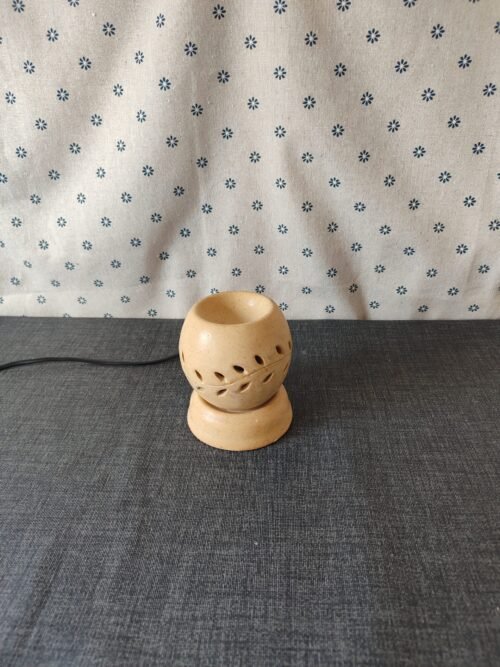 Lámpara de cerámica decorativa con diseño de hojas, sobre una superficie gris, con fondo de tela blanca estampada. Ideal para aromaterapia y decoración del hogar.