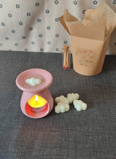 Humificador de cerámica rosa con vela encendida, rodeado de formas de nubes de cera y una caja de cartón decorativa con cerillas. Ideal para aromaterapia y decoración del hogar.