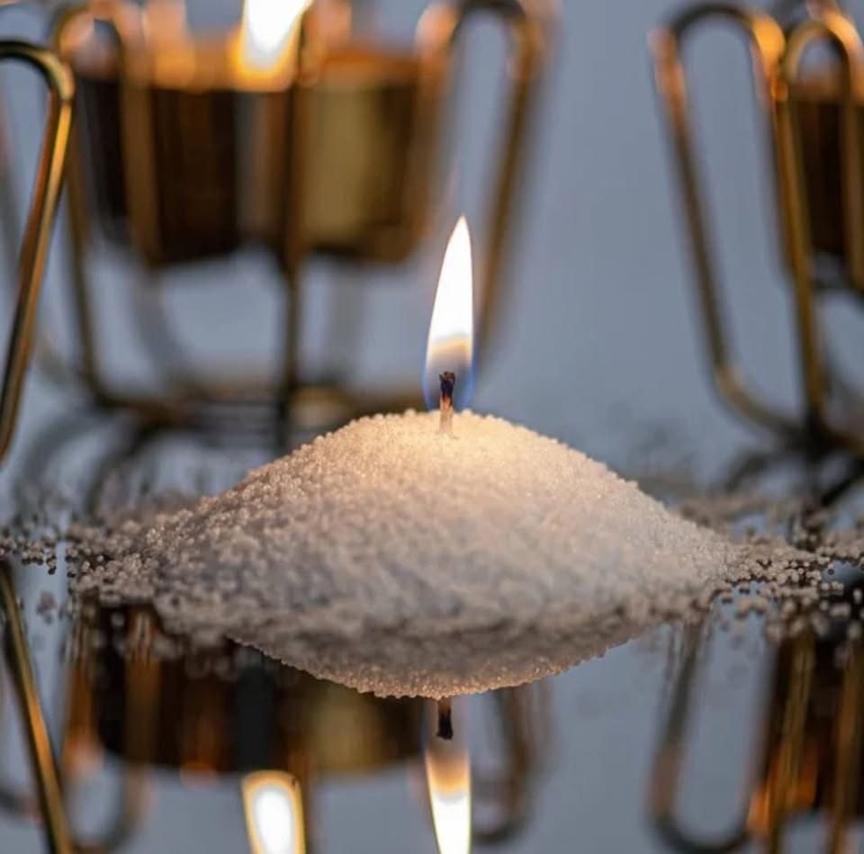 Luz de una vela encendida sobre una montaña de sal, reflejándose en una superficie brillante, con candelabros dorados desenfocados al fondo.